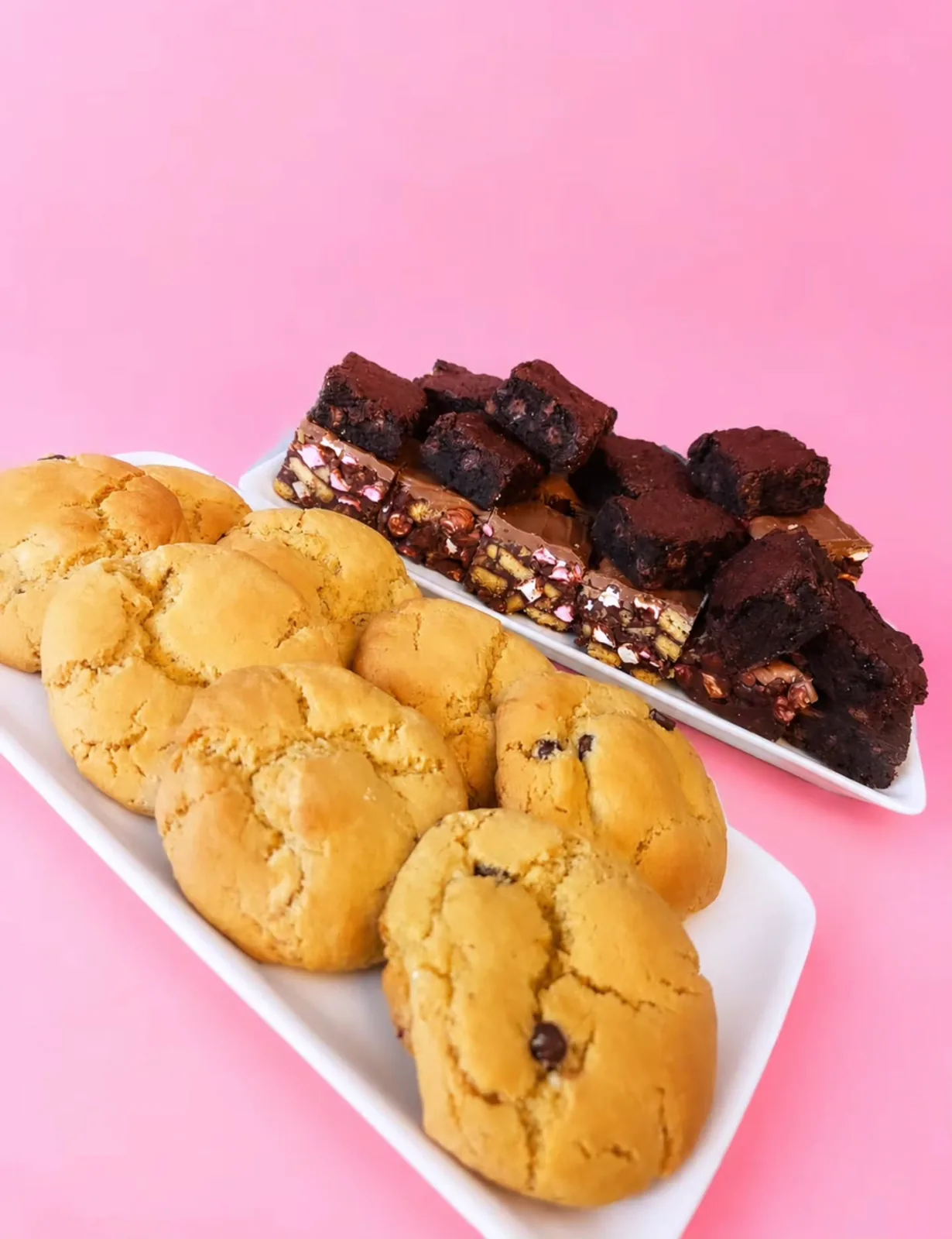 Cookies on a white platter with brownies and a chocolate traybake behind, on a pink backdrop