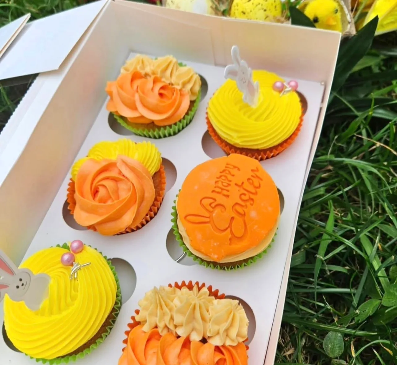 Box of yellow and orange Easter cupcakes with bunny toppers and a Happy Easter disc, on the grass