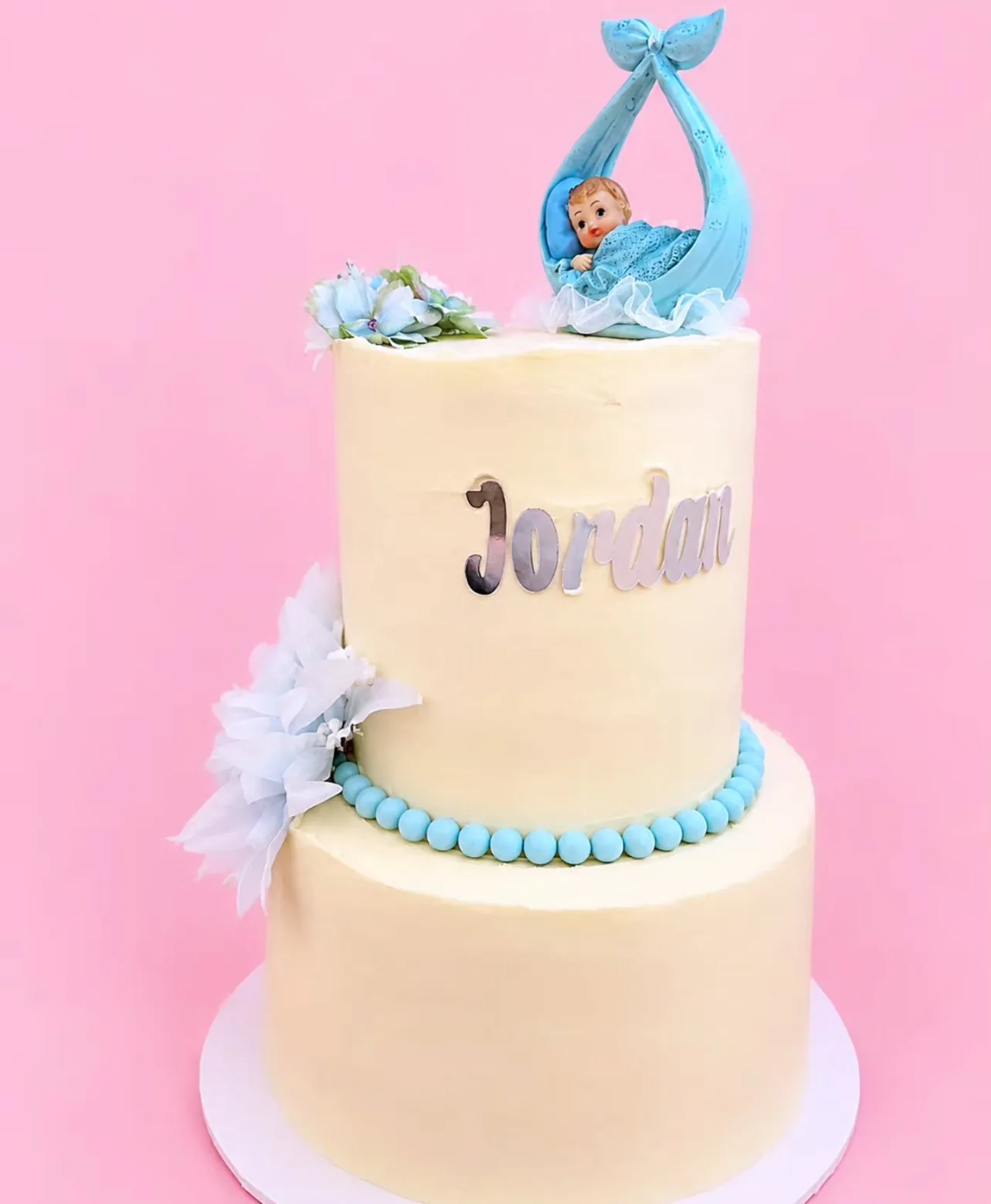 Two-tier cream cake with blue pearls, a silver Jordan topper and a baby in a blue sling on top